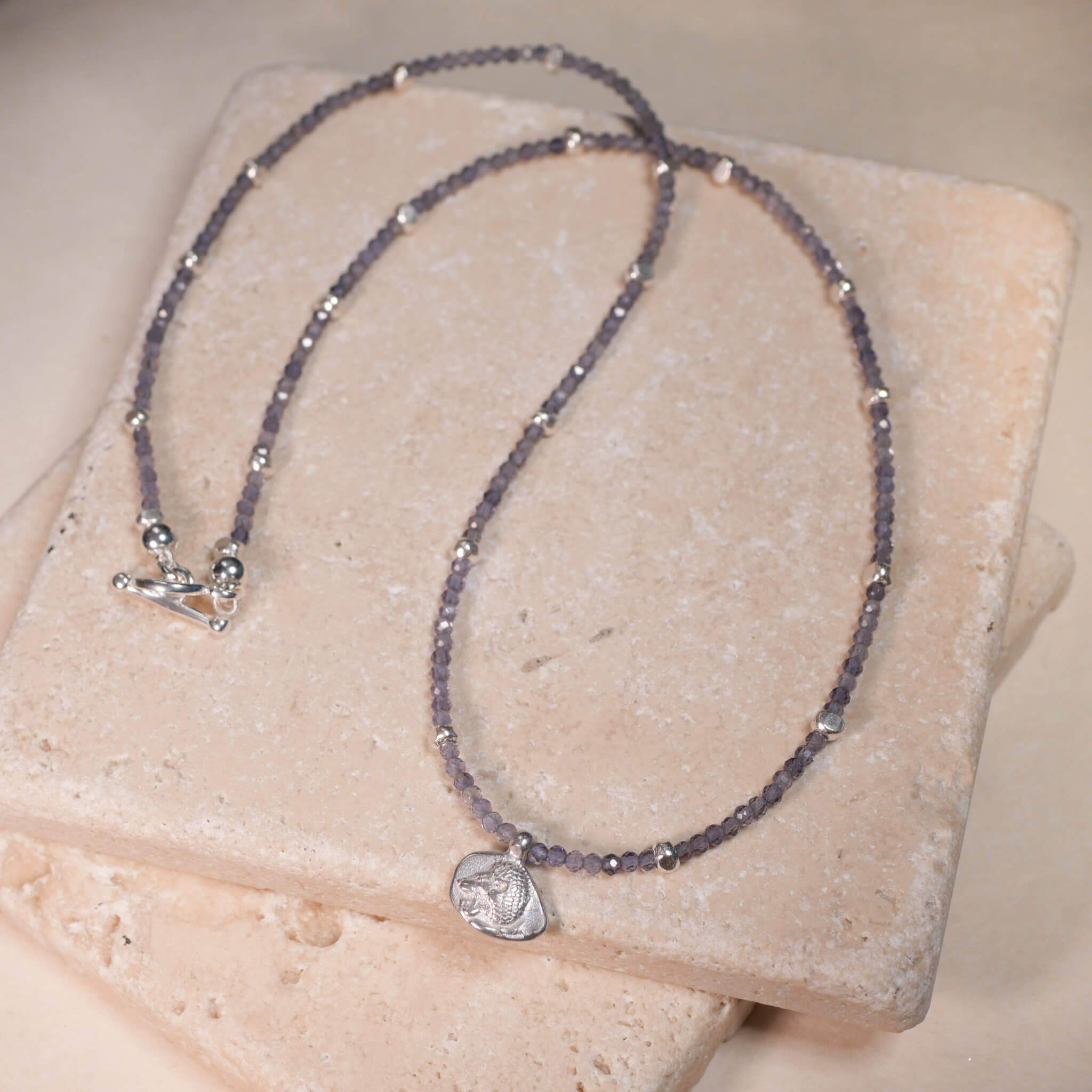 Kette mit der Münze Leona aus rauchblauen iolith Edelsteinen. Der Münzanhänger hat zwei Seiten eine mit Stern und eine mit Löwen. Hier ist der Löwenkopf zu sehen. aus Sterling Silber. True Nuggets. Diese Münze ist eine Nachprägung einer antiken griechischen Münze um ca. 511 v. Chr.
